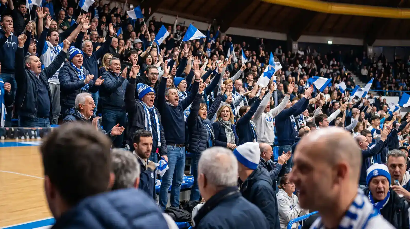 Tifosi italiani di pallacanestro festeggiano in piedi sulle tribune di un palazzetto durante un canestro decisivo
